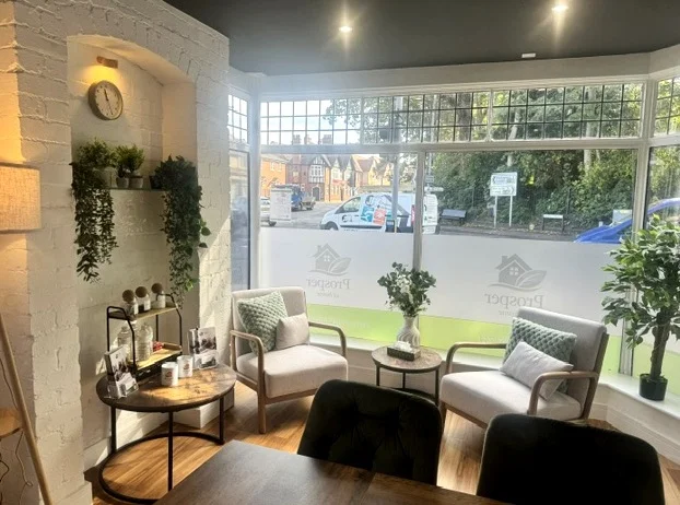 Inside the Prosper at Home office: two armchairs with green cushions by a large window, a small round table with flowers, and plants on a white brick wall